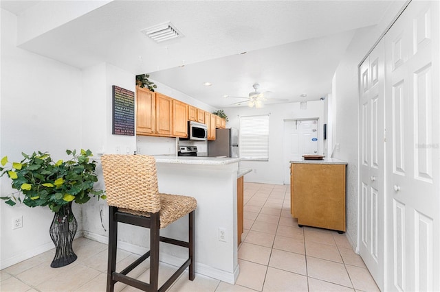 kitchen featuring ceiling fan, light tile patterned floors, light brown cabinetry, appliances with stainless steel finishes, and a breakfast bar area