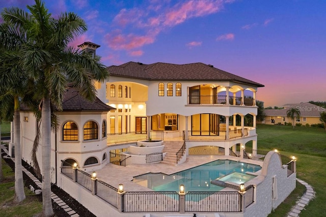 back house at dusk featuring a balcony, a yard, a patio, and a swimming pool with hot tub