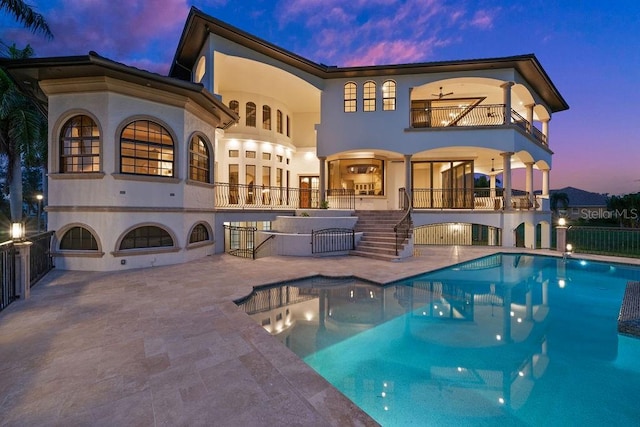 back of property at dusk with a patio, a balcony, an outdoor pool, ceiling fan, and stairs