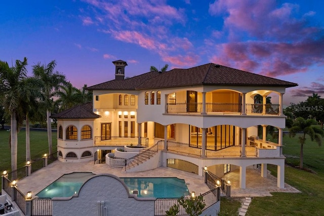 back house at dusk featuring a patio area, a balcony, and a lawn