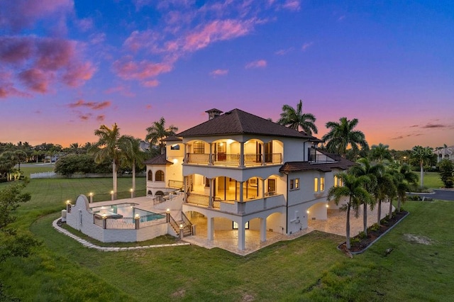 back house at dusk featuring a balcony, a yard, and a pool with hot tub