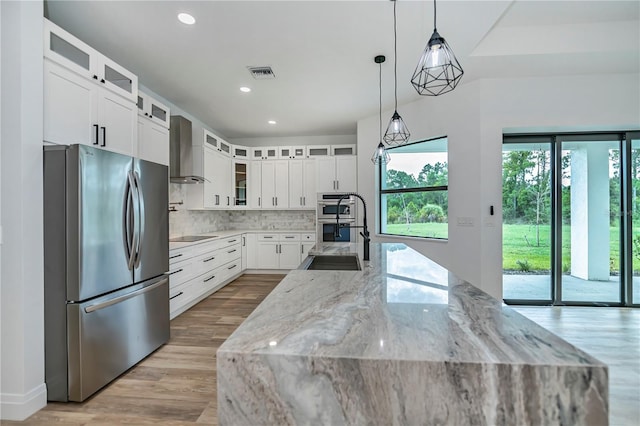 kitchen featuring decorative light fixtures, wall chimney range hood, white cabinetry, light stone countertops, and stainless steel appliances