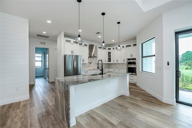 kitchen with white cabinets, appliances with stainless steel finishes, wall chimney range hood, a kitchen island with sink, and light stone counters