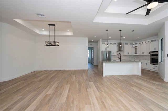 kitchen featuring decorative light fixtures, a raised ceiling, white cabinetry, appliances with stainless steel finishes, and an island with sink