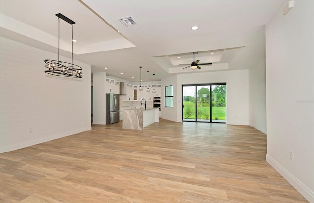 unfurnished living room with ceiling fan with notable chandelier, sink, light hardwood / wood-style flooring, and a raised ceiling