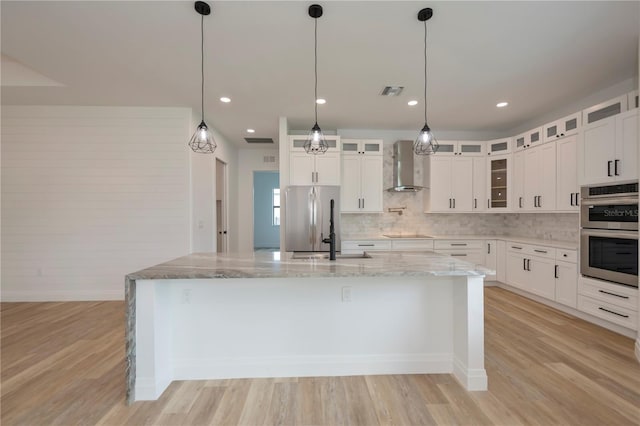 kitchen featuring appliances with stainless steel finishes, wall chimney range hood, an island with sink, light wood-type flooring, and light stone counters