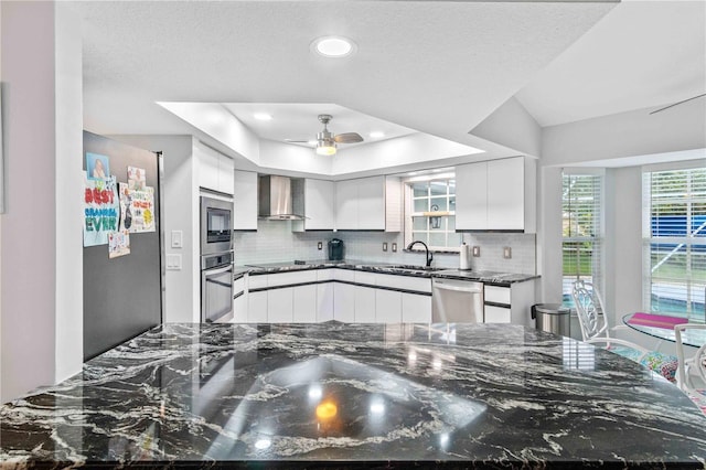 kitchen featuring dark stone countertops, wall chimney range hood, a tray ceiling, white cabinetry, and appliances with stainless steel finishes