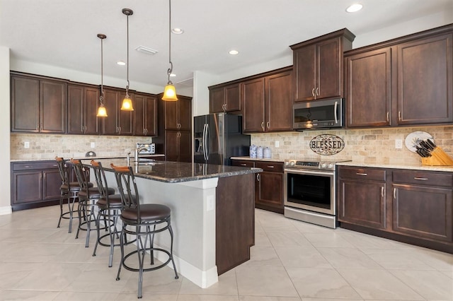 kitchen featuring appliances with stainless steel finishes, an island with sink, dark stone counters, hanging light fixtures, and dark brown cabinets