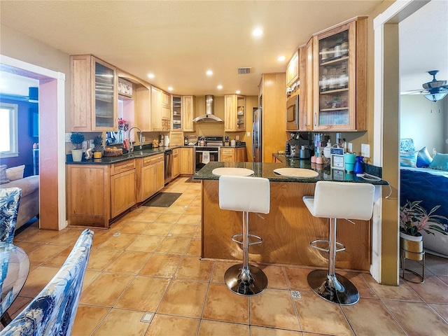 kitchen featuring a breakfast bar area, glass insert cabinets, appliances with stainless steel finishes, wall chimney range hood, and a peninsula