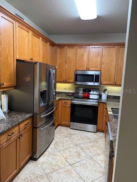 kitchen featuring light tile patterned floors, a textured ceiling, appliances with stainless steel finishes, brown cabinets, and dark stone countertops