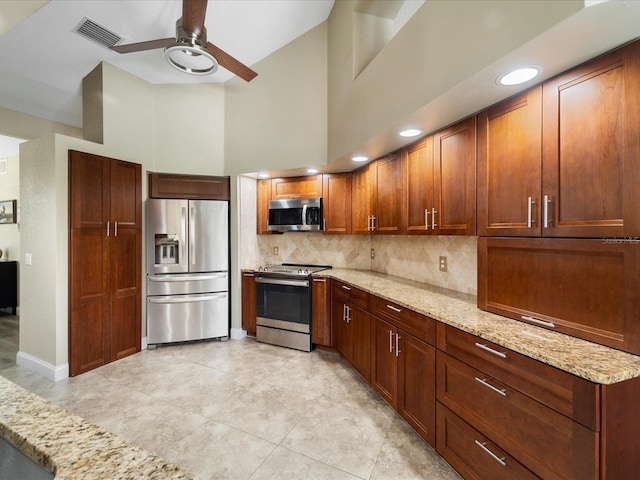 kitchen featuring appliances with stainless steel finishes, a towering ceiling, backsplash, ceiling fan, and light stone countertops