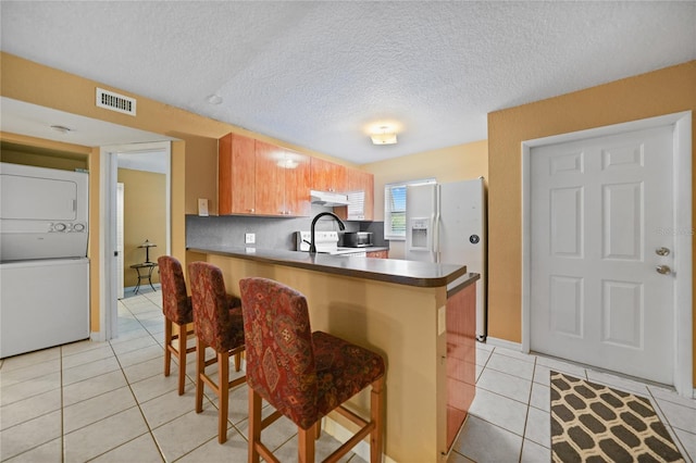 kitchen featuring light tile patterned floors, white appliances, a breakfast bar area, stacked washer and clothes dryer, and kitchen peninsula