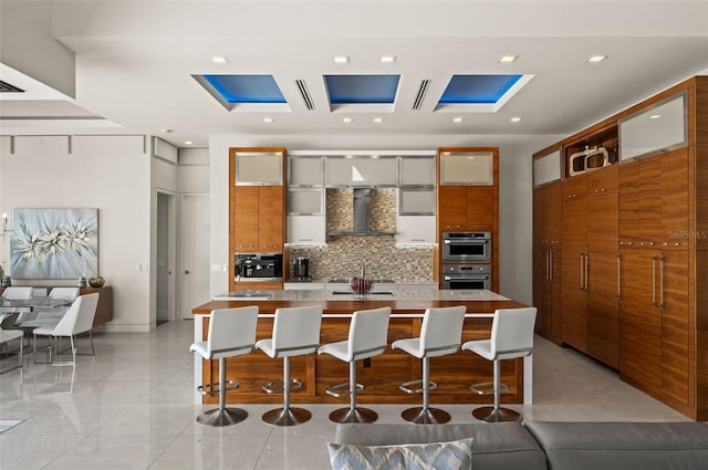 kitchen featuring backsplash, wall chimney range hood, stainless steel double oven, a skylight, and light tile patterned floors