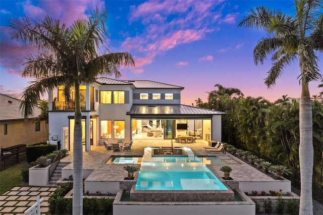 back house at dusk with an outdoor living space, a pool with hot tub, a balcony, and a patio