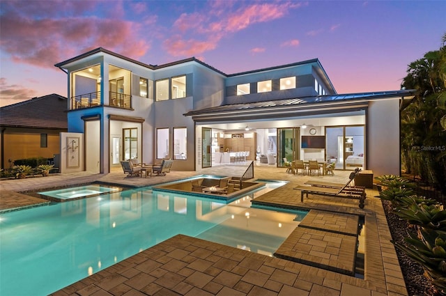 back of house at dusk featuring a patio area, stucco siding, a pool with connected hot tub, and a balcony