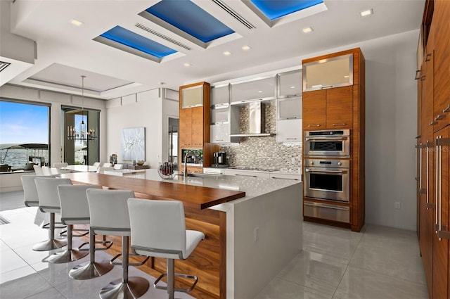 kitchen with a warming drawer, light tile patterned flooring, double oven, wall chimney range hood, and backsplash