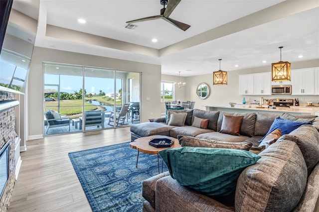 living room with a fireplace, light wood-type flooring, a tray ceiling, and ceiling fan with notable chandelier