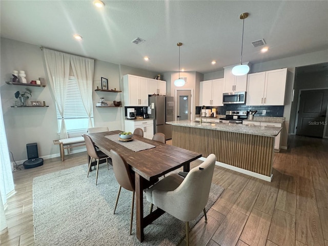 kitchen with appliances with stainless steel finishes, an island with sink, white cabinets, hanging light fixtures, and light stone counters