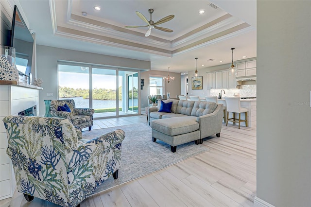 living room featuring a water view, ornamental molding, a tray ceiling, light hardwood / wood-style floors, and ceiling fan with notable chandelier