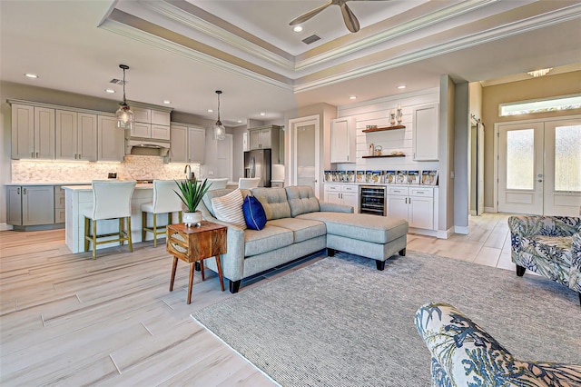 living room with wine cooler, a tray ceiling, light hardwood / wood-style floors, ornamental molding, and french doors