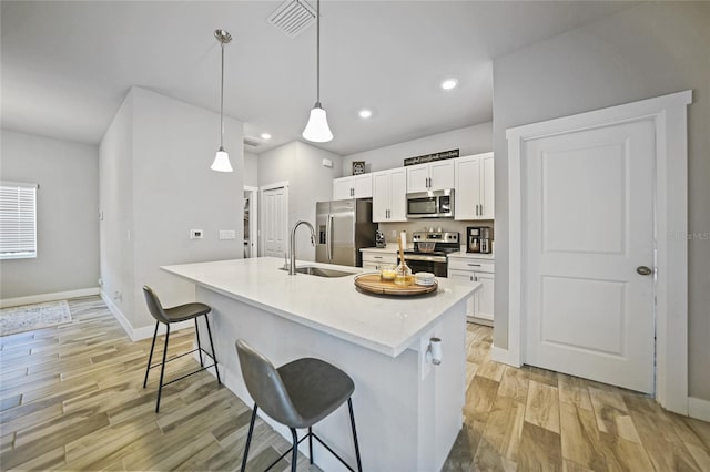 kitchen featuring white cabinets, light hardwood / wood-style floors, appliances with stainless steel finishes, decorative light fixtures, and a center island with sink