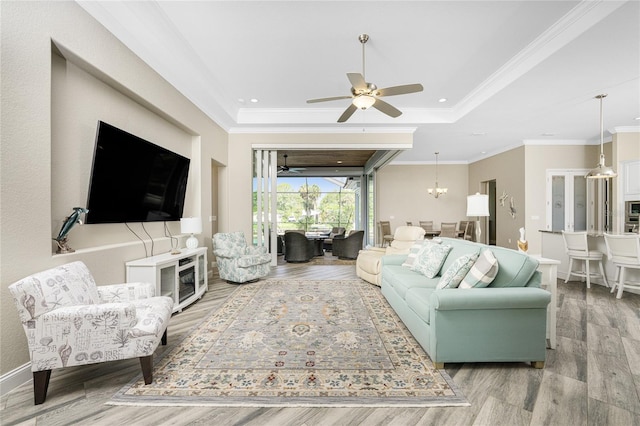 living room featuring a raised ceiling, ornamental molding, ceiling fan with notable chandelier, and light wood-type flooring