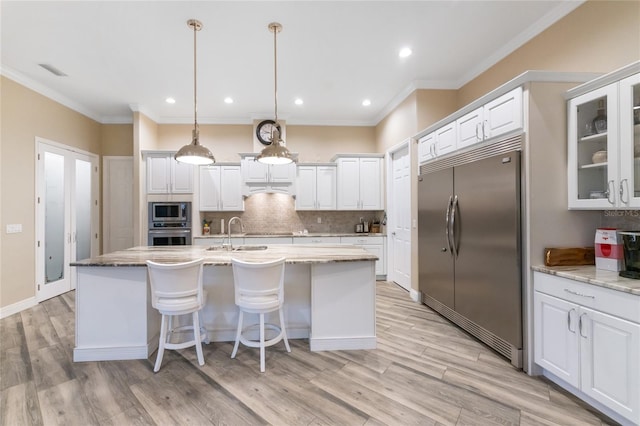 kitchen with pendant lighting, built in appliances, light stone countertops, an island with sink, and white cabinets