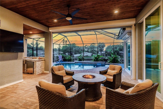 patio terrace at dusk featuring area for grilling, a lanai, a bar, ceiling fan, and an outdoor living space with a fire pit