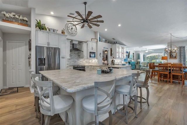 kitchen featuring wall chimney range hood, light wood-type flooring, ceiling fan with notable chandelier, vaulted ceiling, and stainless steel appliances