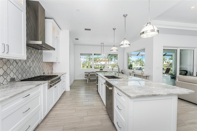 kitchen featuring pendant lighting, an island with sink, sink, wall chimney range hood, and white cabinets