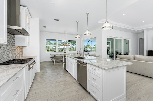 kitchen with stainless steel appliances, wall chimney range hood, white cabinets, and a kitchen island with sink