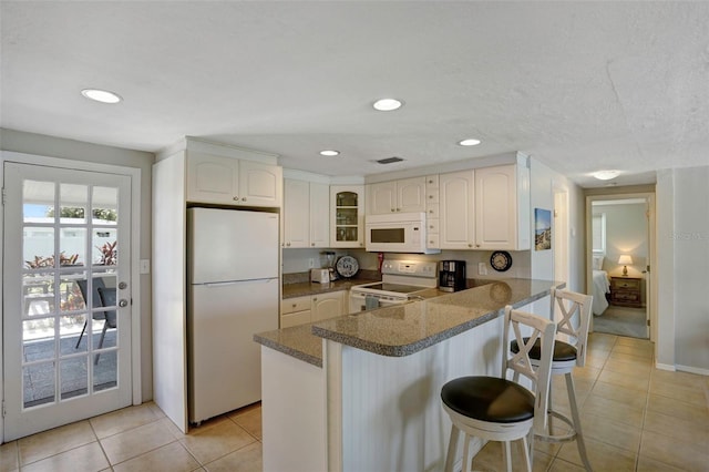 kitchen with light tile patterned flooring, white cabinets, kitchen peninsula, a kitchen bar, and white appliances