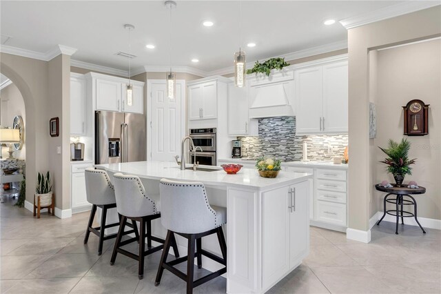 kitchen with a breakfast bar area, premium range hood, a kitchen island with sink, stainless steel appliances, and white cabinets