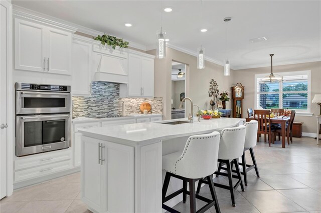 kitchen with white cabinets, stainless steel double oven, an island with sink, and custom exhaust hood