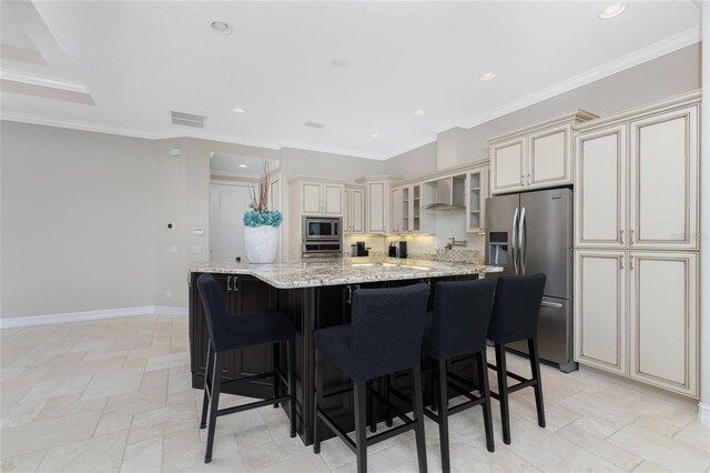 kitchen featuring light stone counters, wall chimney exhaust hood, cream cabinetry, stainless steel appliances, and decorative backsplash