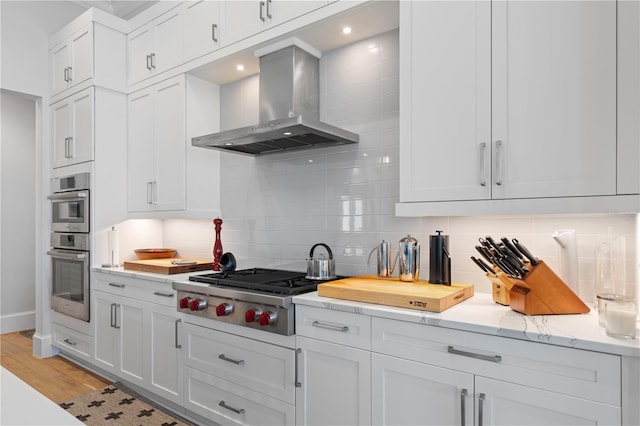 kitchen with appliances with stainless steel finishes, tasteful backsplash, white cabinetry, wall chimney range hood, and light wood-type flooring