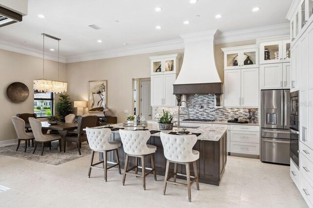 kitchen featuring decorative light fixtures, stainless steel appliances, premium range hood, an island with sink, and white cabinets