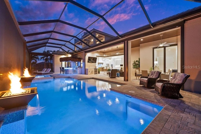 pool at dusk featuring glass enclosure, ceiling fan, a patio, and a fire pit