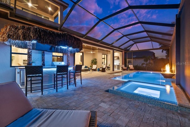 pool at dusk featuring an outdoor bar, an in ground hot tub, ceiling fan, a patio, and glass enclosure