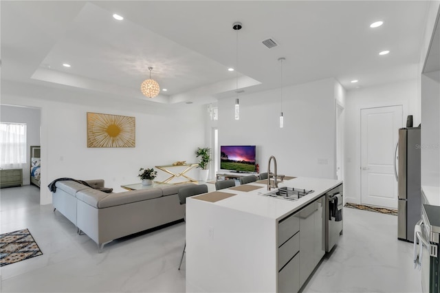 kitchen featuring a raised ceiling, an island with sink, sink, and pendant lighting