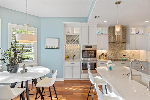 kitchen featuring light hardwood / wood-style floors, white cabinets, pendant lighting, stainless steel double oven, and wall chimney range hood