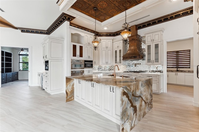 kitchen with light stone counters, sink, decorative backsplash, a center island with sink, and premium range hood