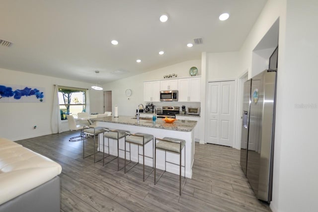 kitchen with white cabinets, vaulted ceiling, a center island with sink, stainless steel appliances, and light hardwood / wood-style floors