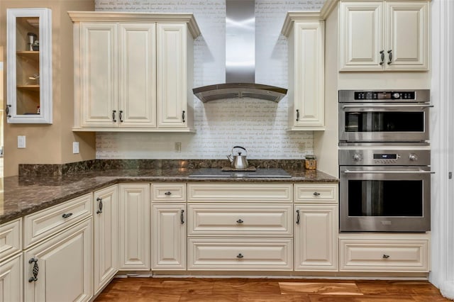 kitchen with black electric cooktop, double oven, wall chimney range hood, cream cabinets, and dark stone countertops