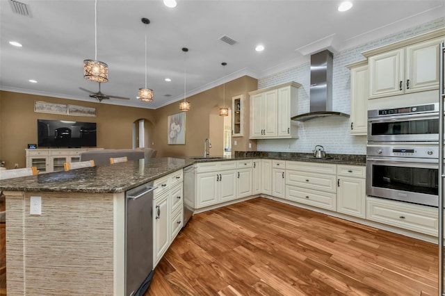 kitchen with kitchen peninsula, cream cabinetry, hanging light fixtures, and wall chimney range hood