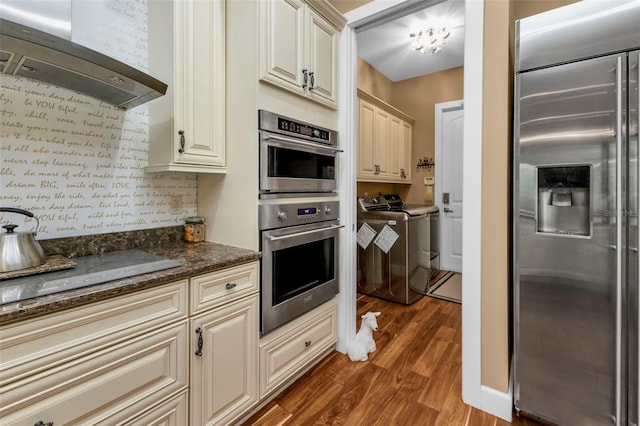 kitchen with ventilation hood, dark stone counters, cream cabinetry, washer and clothes dryer, and appliances with stainless steel finishes