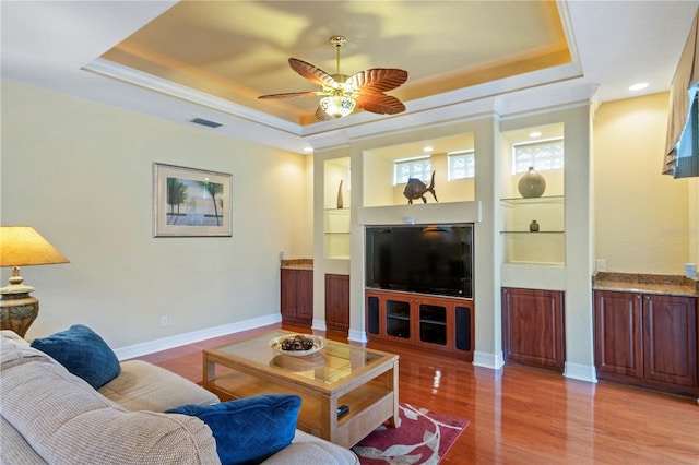 living room with built in shelves, light hardwood / wood-style flooring, a raised ceiling, crown molding, and ceiling fan