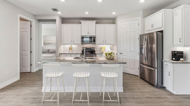 kitchen with white cabinets, light stone counters, stainless steel appliances, and an island with sink