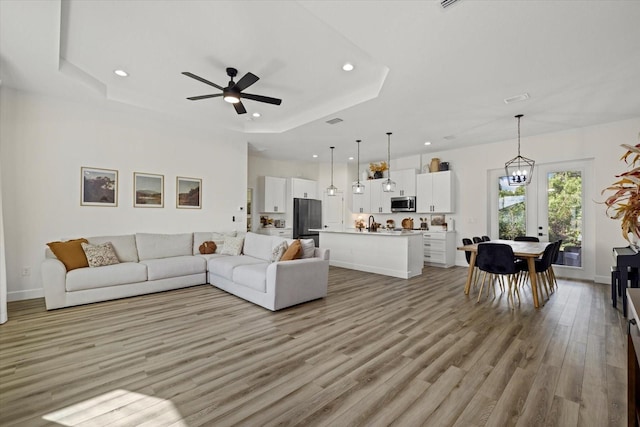 living room featuring ceiling fan with notable chandelier, sink, light wood-type flooring, and a raised ceiling