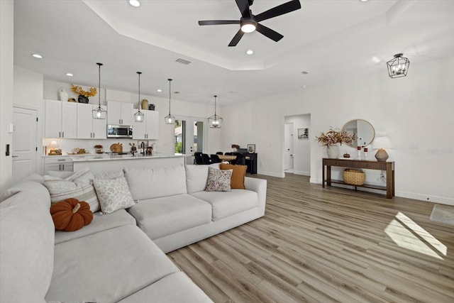 living room with ceiling fan with notable chandelier, a tray ceiling, and light hardwood / wood-style flooring